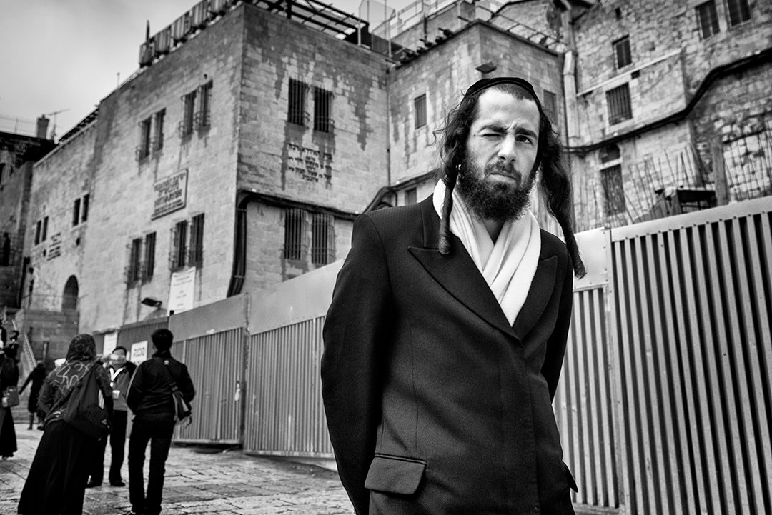 A bearded man walking in front of stone buildings.